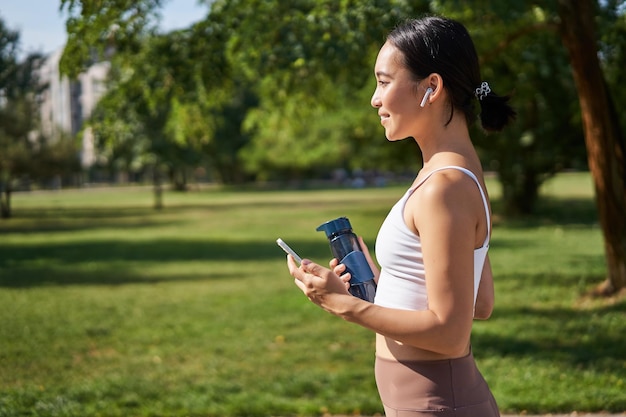 Actief aziatisch meisje in fitness kleding training in park wandelen in sportkleding met smartphone en water