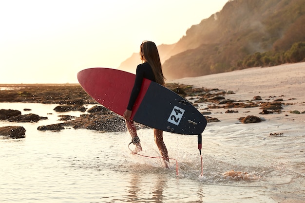 Actief sport- en levensstijlconcept. Boardsurfer in beweging, draagt rode surfplank, rent het water in, geniet van vrije tijd om te surfen