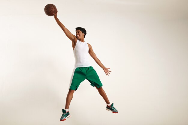 Actiefoto van een gelukkige jonge zwarte atleet die een wit overhemd en een groene korte broek draagt die hoog springt om een vintage basketbal op wit te grijpen