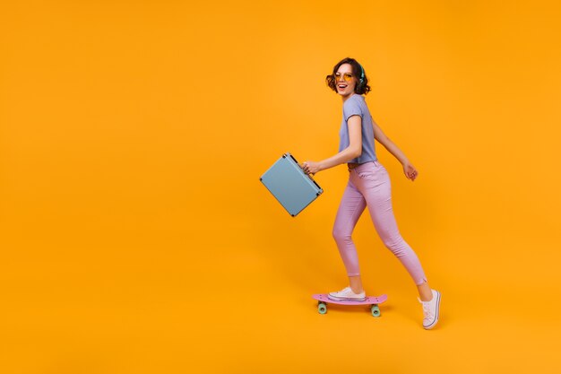 Actieve blanke vrouw met valise schaatsen. Binnen schot van prachtig krullend meisje dat zich op longboard bevindt.