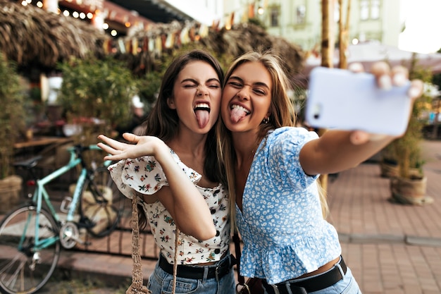 Actieve jonge meisjes in stijlvolle blouses trekken grappige gezichten, tonen tongen en nemen selfie