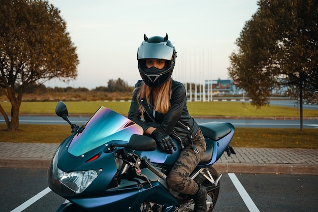 Actieve levensstijl, extreme en adrenaline-concept. Outdoor Portret van modieuze jonge blonde vrouw kaki jeans, veiligheidshelm, zwart lederen handschoenen en jas poseren op motorfiets