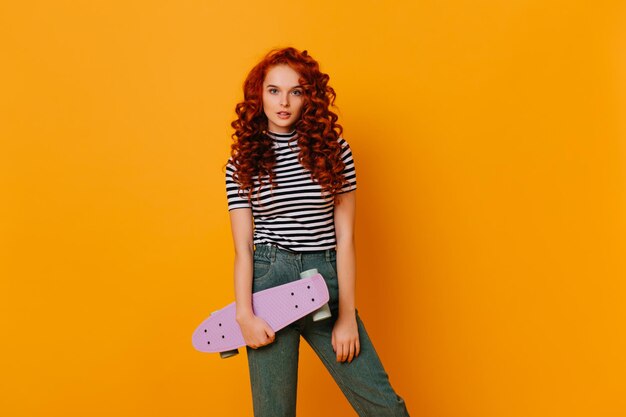 Actieve roodharige meisje in stijlvolle denim broek en top poseren met skateboard op oranje achtergrond