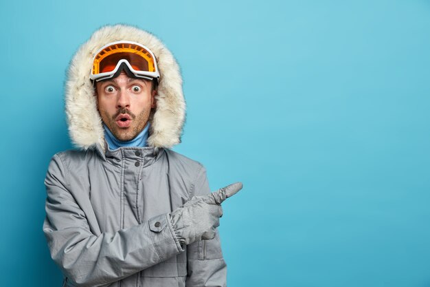 Actieve stomverbaasde man traint in de winterbergen gaat snowboarden draagt een grijze jas en een skibril geeft met verbaasde uitdrukking op lege ruimte aan.