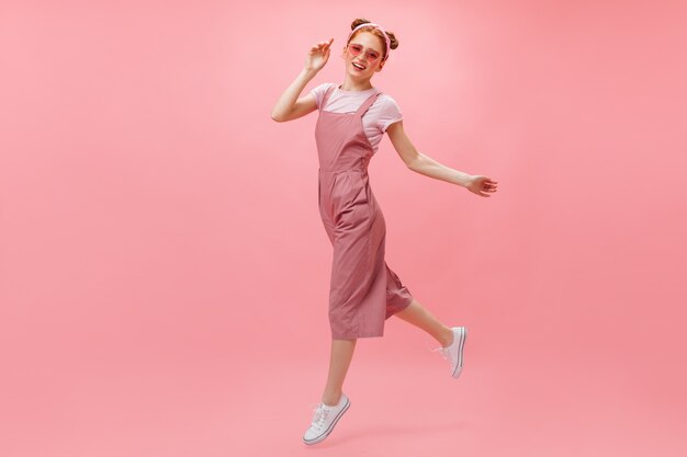 Actieve vrouw in roze jumpsuit, T-shirt en stijlvolle bril beweegt op roze achtergrond.