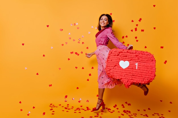 Actieve vrouw in roze kleren die pret hebben. Emotioneel meisje dat met bruin haar op oranje muur springt.