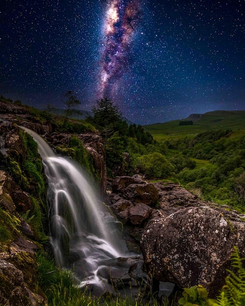 Gratis foto adembenemende scène van een waterval, groene velden, rotsen onder een blauwe lucht met glanzende sterren 's nachts