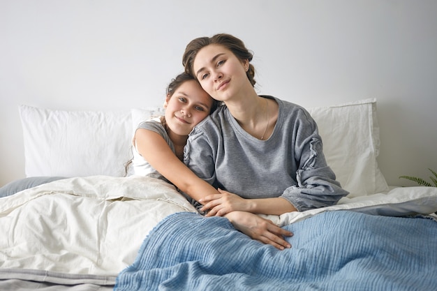 Adorably schattig vrouwelijk kind zit in bed en omhelst haar aantrekkelijke moeder.