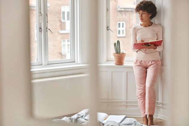 Afbeelding van doordachte donkere huid freelancer maakt aantekeningen in kladblok, draagt nachtkleding