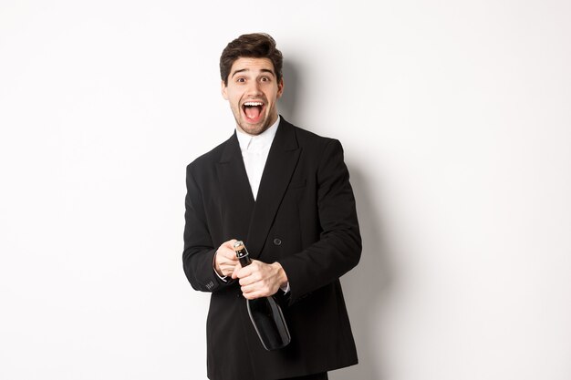 Afbeelding van een aantrekkelijke man in een zwart pak die een feestje viert, het nieuwe jaar viert en een fles champagne opent, die gelukkig staat tegen een witte achtergrond