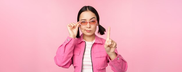 Afbeelding van een Aziatisch meisje met een serieus gezicht dat een uitbranderig gebaar laat zien dat de vinger schudt die wijst op uitdrukkelijke afkeuring die over roze achtergrond staat