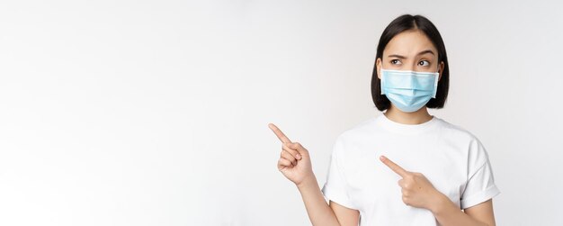 Afbeelding van een aziatische vrouw met een medisch masker van covid die verward naar links kijkt, wijzend naar het logo met een advertentie die op een witte achtergrond staat