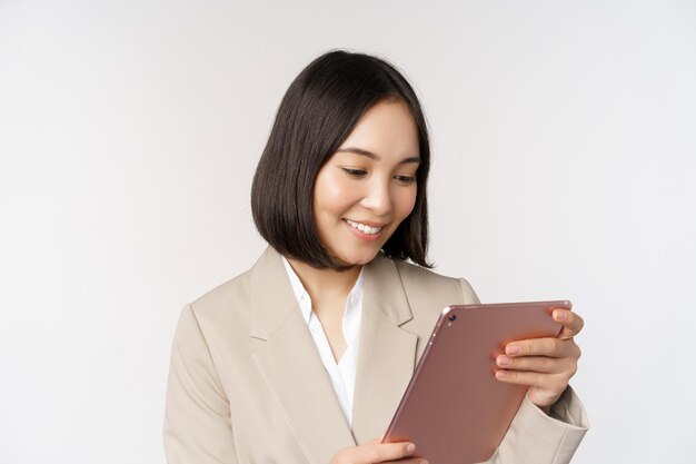 Afbeelding van een Aziatische zakenvrouw die een digitale tablet gebruikt die naar een gadget kijkt en glimlacht terwijl ze tegen een witte achtergrond staat
