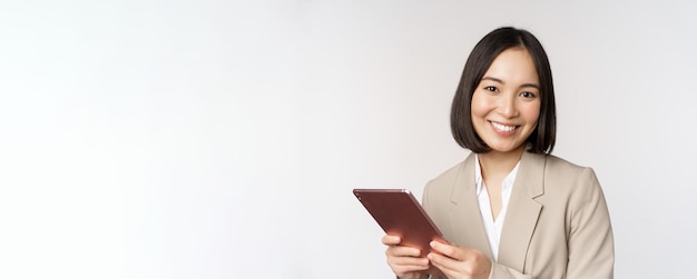 Afbeelding van een Aziatische zakenvrouw die een digitale tablet vasthoudt en glimlacht terwijl ze met een gadget in een pak werkt op een witte achtergrond