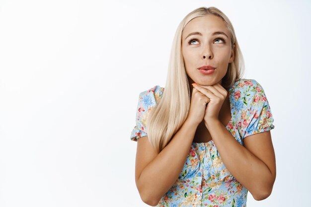 Afbeelding van een dromerige blonde vrouw die opkijkt met een gefascineerde doordachte uitdrukking die iets voorstelt dat in een jurk op een witte achtergrond staat