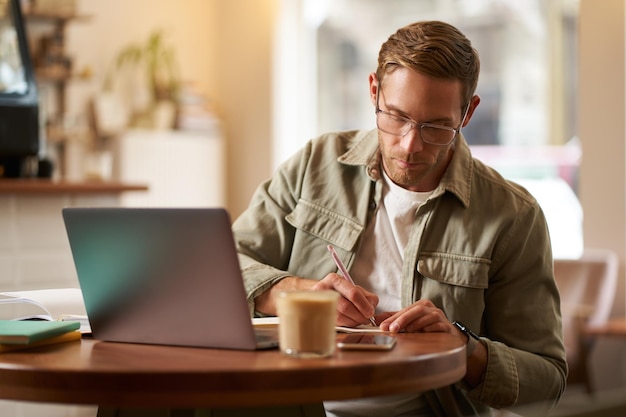 Afbeelding van een gefocuste jonge man met een bril die in een café zit, aantekeningen maakt, studeert, een online cursus volgt