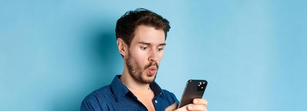 Afbeelding van een geïmponeerde jonge man die naar een smartphone kijkt en wow zegt terwijl hij online aanbiedingen bekijkt