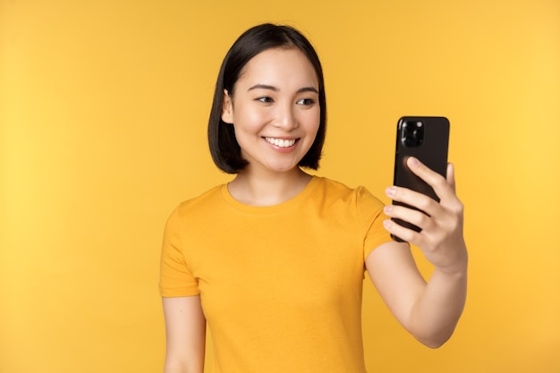 Afbeelding van een gelukkig mooi Aziatisch meisje dat aan het videochatten is op een smartphone-applicatie die tegen een gele achtergrond staat