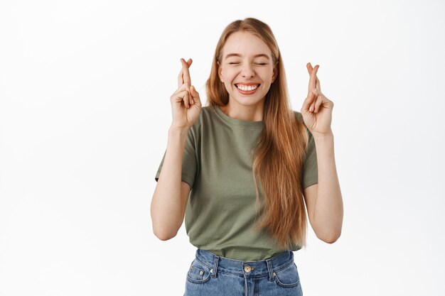 Afbeelding van een gelukkig optimistisch blond meisje, sluit de ogen en kruis vingers voor geluk, bidden, wachten op een goed teken van fortuin, smeken om een wens die uitkomt, staande over een witte muur