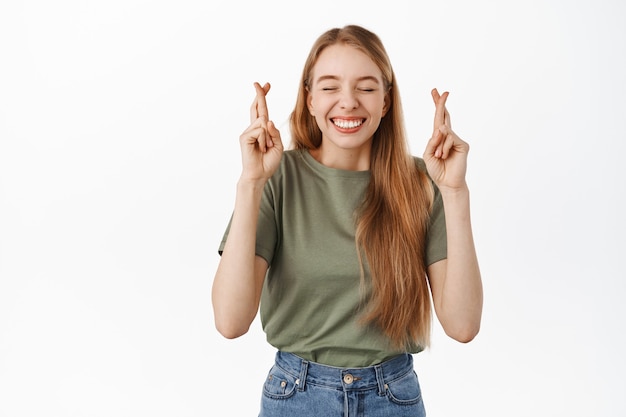 Afbeelding van een gelukkig optimistisch blond meisje, sluit de ogen en kruis vingers voor geluk, bidden, wachten op een goed teken van fortuin, smeken om een wens die uitkomt, staande over een witte muur