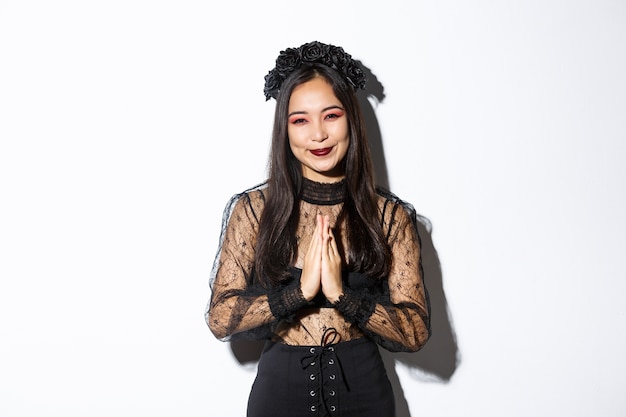 Afbeelding van een gelukkige en dankbare jonge Aziatische vrouw in gotische kanten jurk gesp handen samen om dank u te zeggen, glimlachend dankbaar en permanent op witte achtergrond.