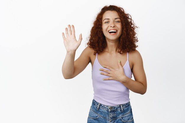 Afbeelding van een gelukkige jonge vrouw steekt haar hand op en lacht, groet, stelt zichzelf voor, naam, belooft met vrolijke glimlach, witte achtergrond.