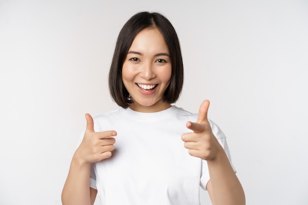 Afbeelding van een glimlachend Aziatisch meisje dat met de vingers naar de camera wijst en ervoor kiest om je uit te nodigen om in een t-shirt op een witte achtergrond te staan