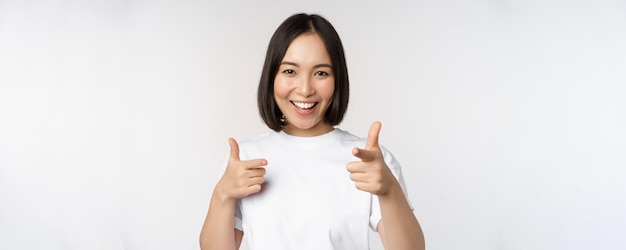 Afbeelding van een glimlachend Aziatisch meisje dat met de vingers naar de camera wijst en ervoor kiest om je uit te nodigen om in een t-shirt op een witte achtergrond te staan