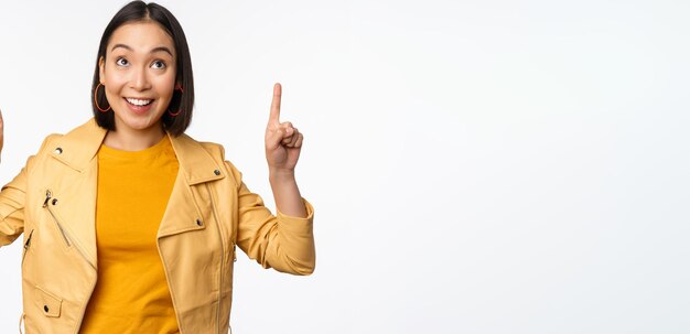 Afbeelding van een glimlachende Aziatische brunette vrouw die met haar vingers omhoog wijst en reclame toont met een blij gezicht dat zich voordeed tegen een witte achtergrond