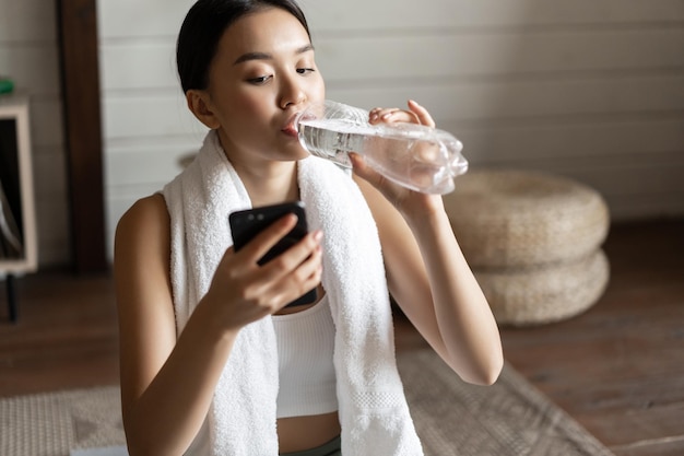 Afbeelding van een jong aziatisch fitnessmeisje met een geschikte lichaamshanddoek om de nek gewikkeld die naar een mobiele telefoon kijkt...