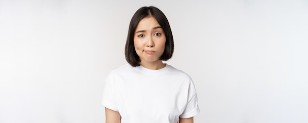 Afbeelding van een jonge Aziatische brunette vrouw in een witte t-shirt grijns en kijk met twijfel aarzelend tegen een witte achtergrond Kopieer de ruimte