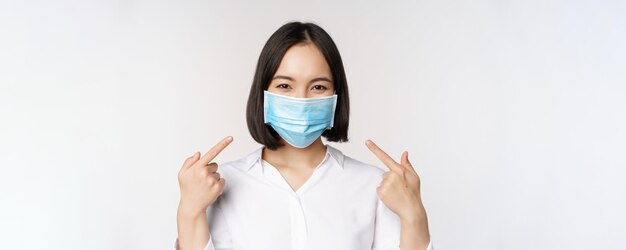 Afbeelding van een jonge aziatische vrouw die naar zichzelf wijst terwijl ze een medisch gezichtsmasker draagt, een concept van covid19-bescherming die op een witte achtergrond staat