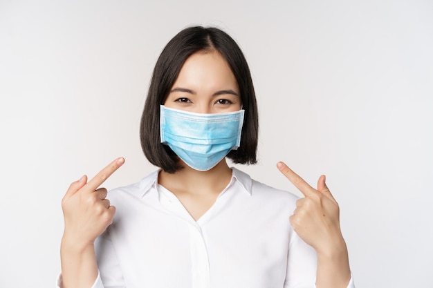 Afbeelding van een jonge aziatische vrouw die naar zichzelf wijst terwijl ze een medisch gezichtsmasker draagt, een concept van covid19-bescherming die op een witte achtergrond staat