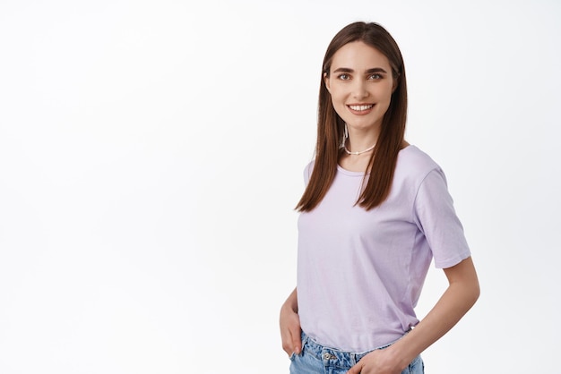 Afbeelding van een jonge moderne vrouw in een t-shirt die naar de camera kijkt met een zelfverzekerde glimlach, hand in hand in jeanszakken, een casual pose van een ontspannen persoon op een witte achtergrond