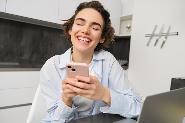 Afbeelding van een jonge vrouw die haar smartphone thuis gebruikt. Meisje zit met mobiele telefoon in de keuken en glimlacht