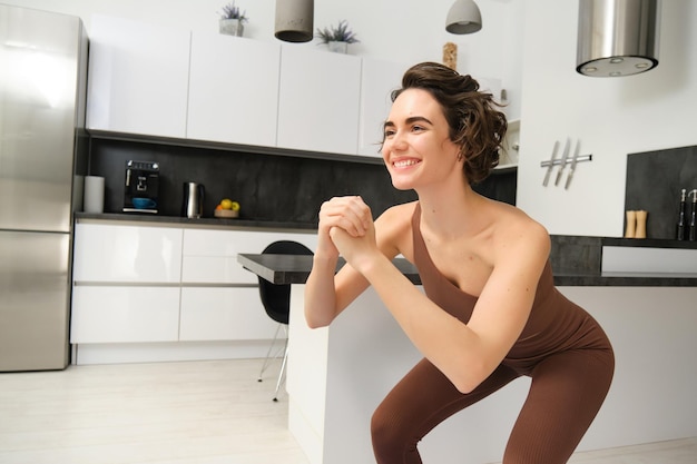 Afbeelding van een jonge vrouw die thuis squats doet, oefent op een rubberen mat in een heldere kamer binnen en draagt activiteit