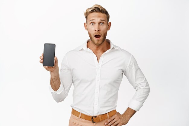 Afbeelding van een knappe blonde man die een smartphonescherm toont met een verbaasd gezicht, laat de kaak vallen en zegt wow verbaasd over een witte achtergrond