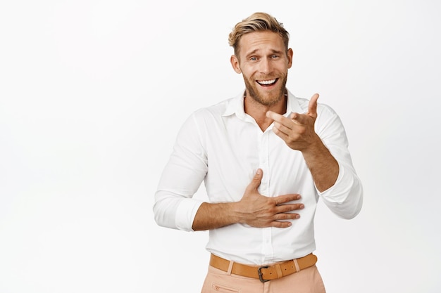 Afbeelding van een knappe blonde man die lacht en met de vinger naar de camera wijst en naar iets grappigs kijkt en grinnikt over een witte achtergrond