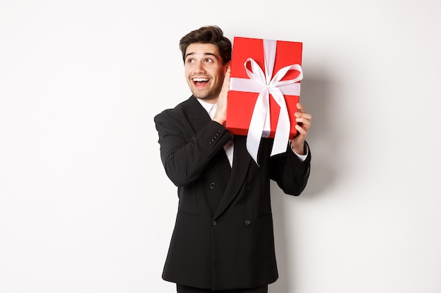 Afbeelding van een knappe dromerige man in een zwart pak, een doos schuddend met een cadeau om zich af te vragen wat erin zit, gelukkig tegen een witte achtergrond.