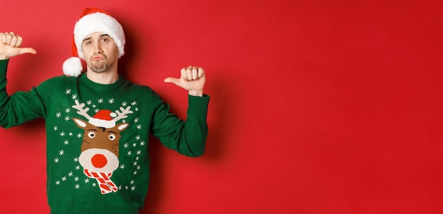 Afbeelding van een knappe en zelfverzekerde jongeman in een groene trui en kerstmuts, wijzend naar zichzelf, kerst vierend, staande op een rode achtergrond.