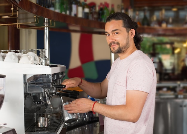 Afbeelding van een knappe jonge glimlachende mannelijke barista die koffiedrank bereidt, met behulp van een koffiemachine van commerciële kwaliteit in de restaurantomgeving.