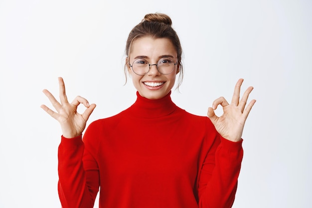 Afbeelding van een knappe jonge vrouw met een bril, tevreden glimlachend, oke tekenen in goedkeuring, iets leuk vinden, goed zeggen, ja gebaar maken op wit