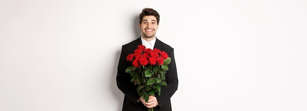 Gratis foto afbeelding van een knappe man in een zwart pak die een boeket rozen vasthoudt en glimlachend tegen een witte rug staat