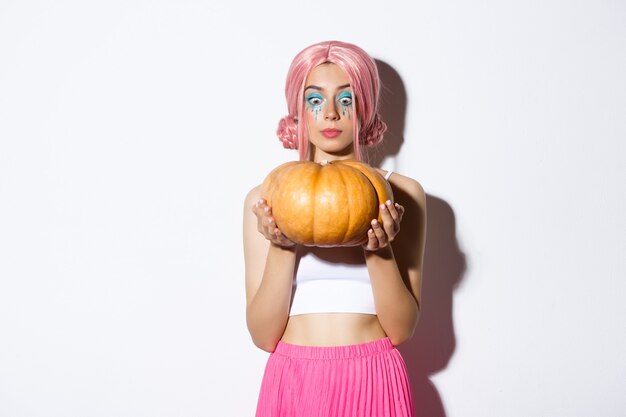 Afbeelding van een mooi meisje met een roze pruik en Halloween-make-up, opgewonden kijkend naar een grote pompoen, staande op een witte achtergrond