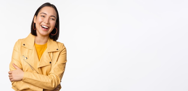 Afbeelding van een mooi modern Aziatisch meisje dat lacht en lacht en gelukkig kijkt naar de camera die in een gele jas staat tegen een witte achtergrond