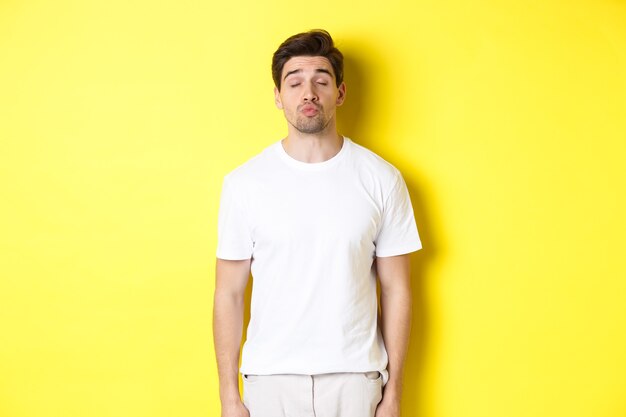 Afbeelding van een mooie man die ogen sluit en lippen tuit, wachtend op een kus, staande in witte kleren tegen een gele achtergrond. Ruimte kopiëren