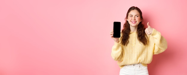 Afbeelding van een mooie vrouw die lacht en opzij kijkt terwijl ze een app beoordeelt met duimen omhoog en een leeg telefoonscherm dat online promo aanbeveelt die tegen een roze achtergrond staat