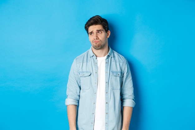 Afbeelding van een onwillige en verdrietige man in een casual outfit die naar links kijkt, fronst en zich overstuur voelt, staande tegen een blauwe achtergrond