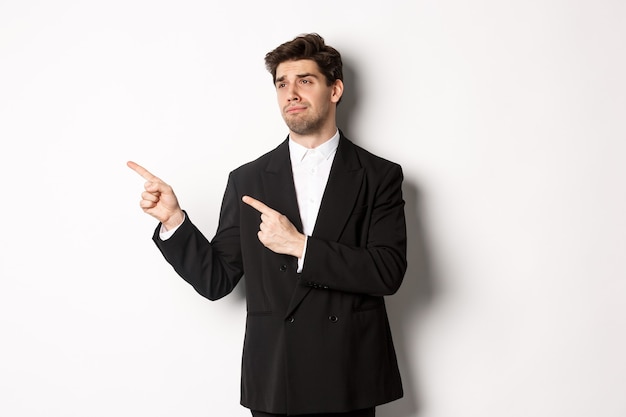 Afbeelding van een overstuur en teleurgestelde knappe man in een formeel pak, wijzend en naar links kijkend met een droevig gezicht, staande op een witte achtergrond