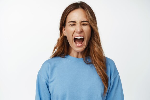 Afbeelding van een pissige vrouw die de druk laat ontsnappen, hardop schreeuwt, met gesloten ogen schreeuwt, in een blauw shirt tegen een witte achtergrond staat